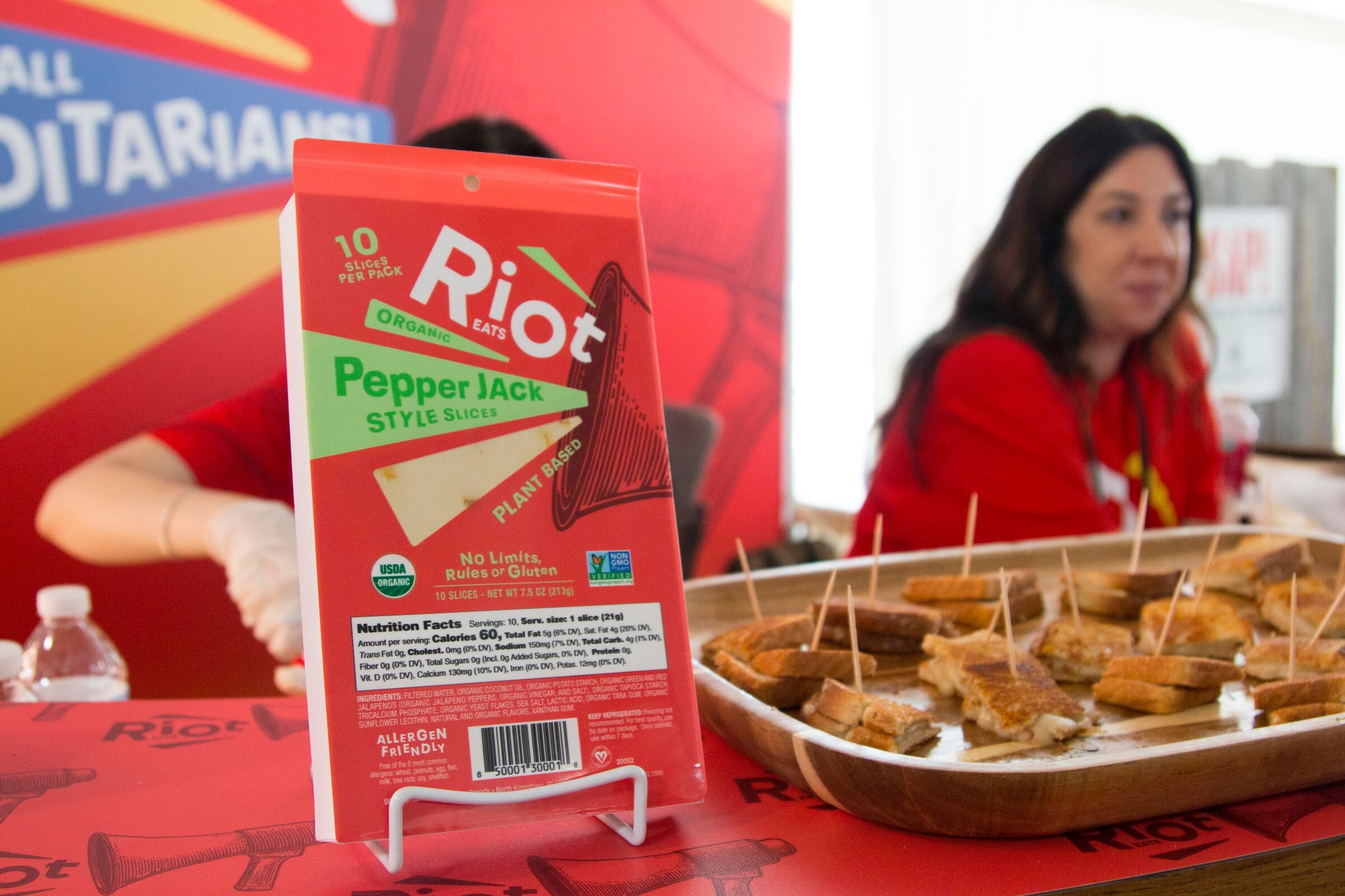 A red Riot Eats package on a stand with a tray of cheese bites and a staffer in red in the background.