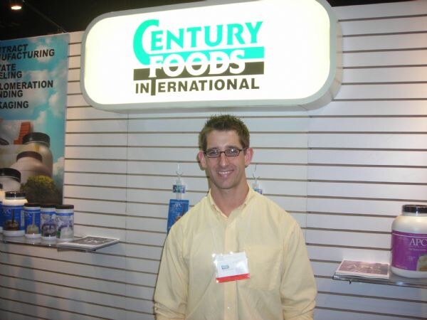 A smiling man with glasses in a yellow shirt stands under a lit Century Foods International sign, with white supplement containers on shelves beside him.