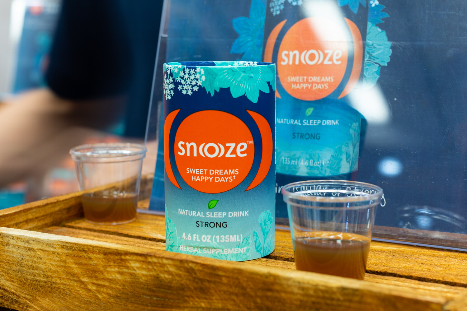 A teal and orange Snooze drink carton on a wooden tray beside small cups of amber liquid.
