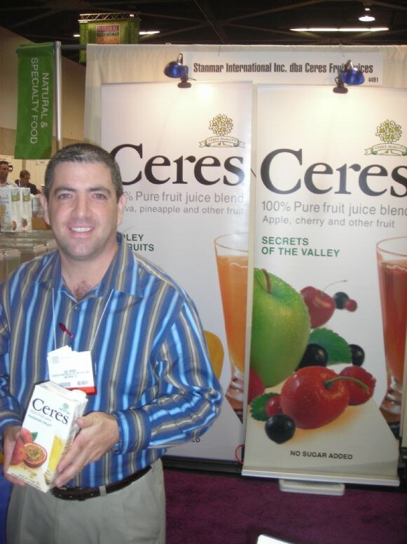 A smiling man in a blue striped shirt holds a carton of Ceres juice in front of Ceres banners for 100% pure fruit juice blends at the Stanmar International Inc. booth.
