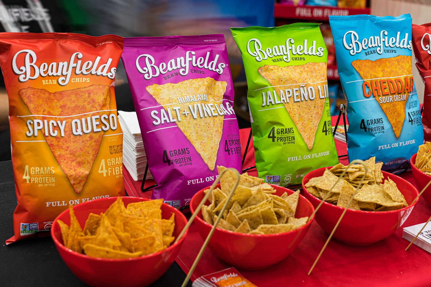 Bags of Beanfields bean chips in Spicy Queso, Himalayan Salt and Vinegar, Jalapeno Lime and Cheddar Sour Cream with red bowls of chips.