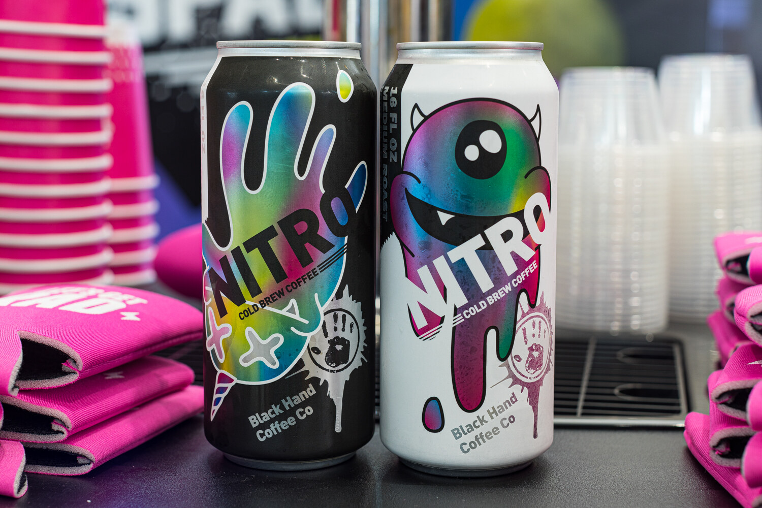 Two colorful cans labeled Nitro Black Hand Coffee Co with pink koozies behind them.