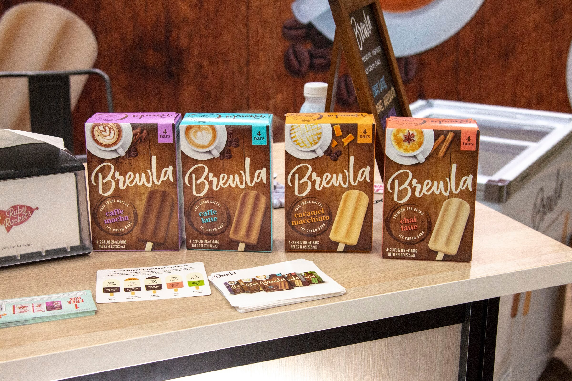 Four Brewla boxes in latte, mocha and other flavors on a wooden counter with a chalkboard sign.