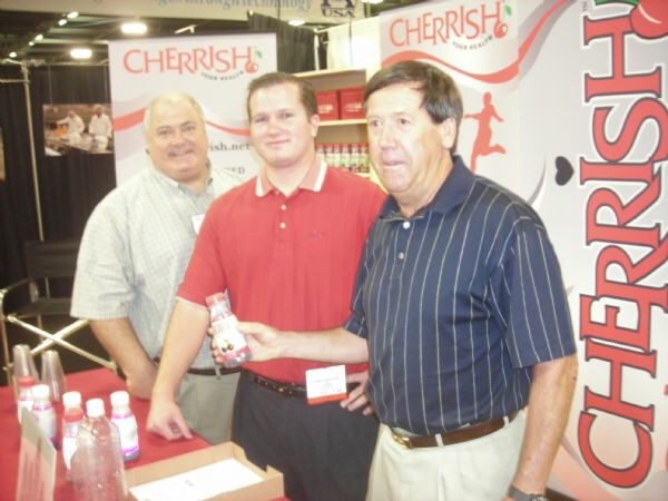 Three men stand together at a booth with Cherrish banners behind them; the man on the right holds a small bottle, and more bottles sit on the red-covered table.