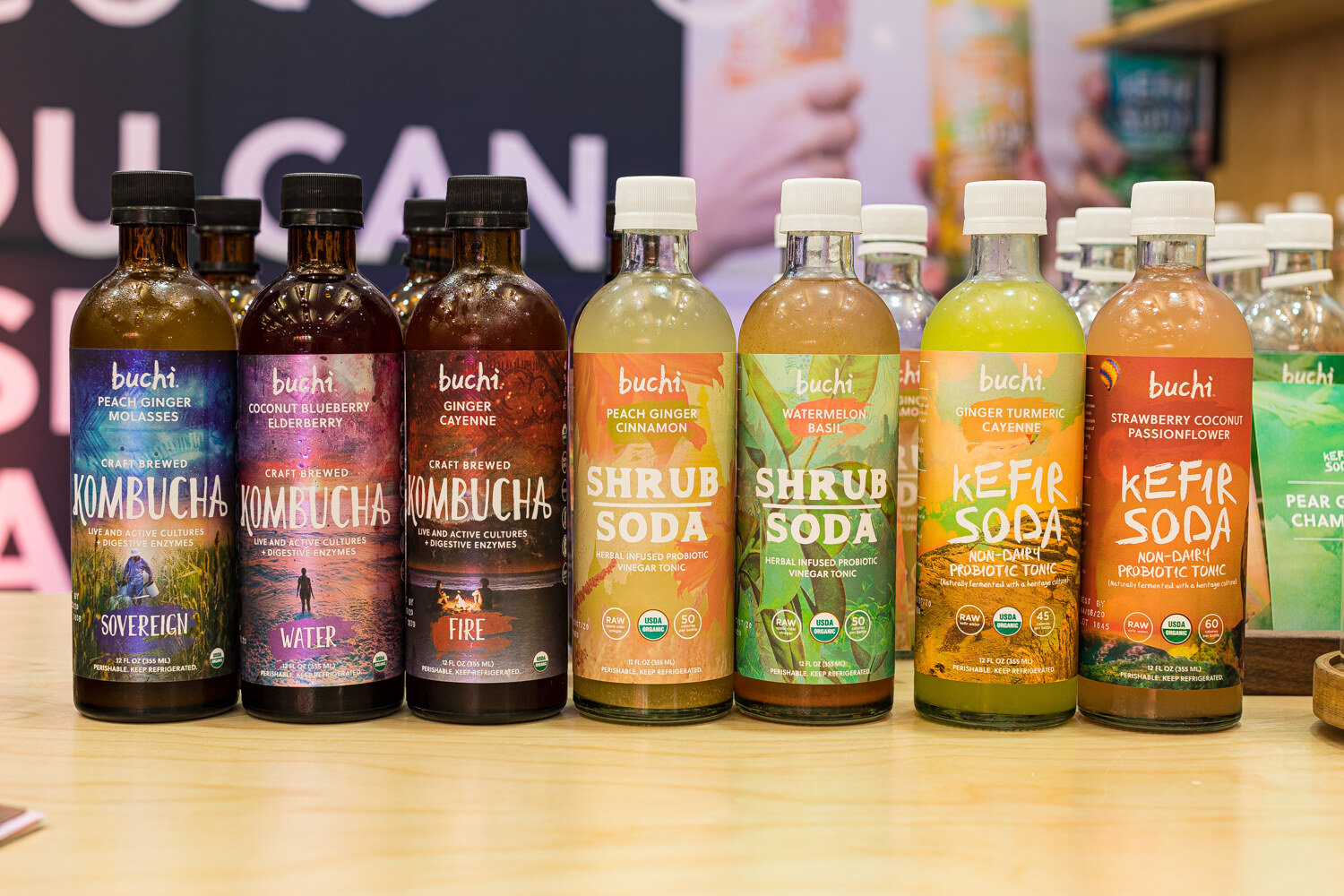 Bottles of Buchi kombucha, shrub soda and kefir soda in a row on a wooden surface.