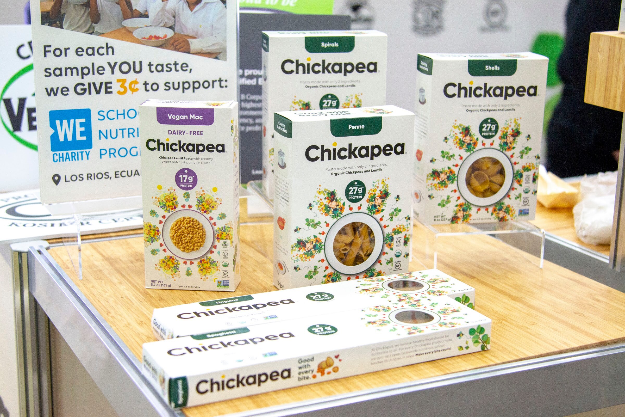 Boxes of Chickapea pasta in several shapes on a table with a sign mentioning WE Charity.