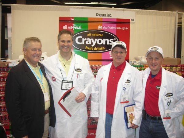Four men pose in front of a Crayons all natural fruit drinks sign; three wear white lab coats with Crayons logos, two of them in caps, and one wears a dark jacket.