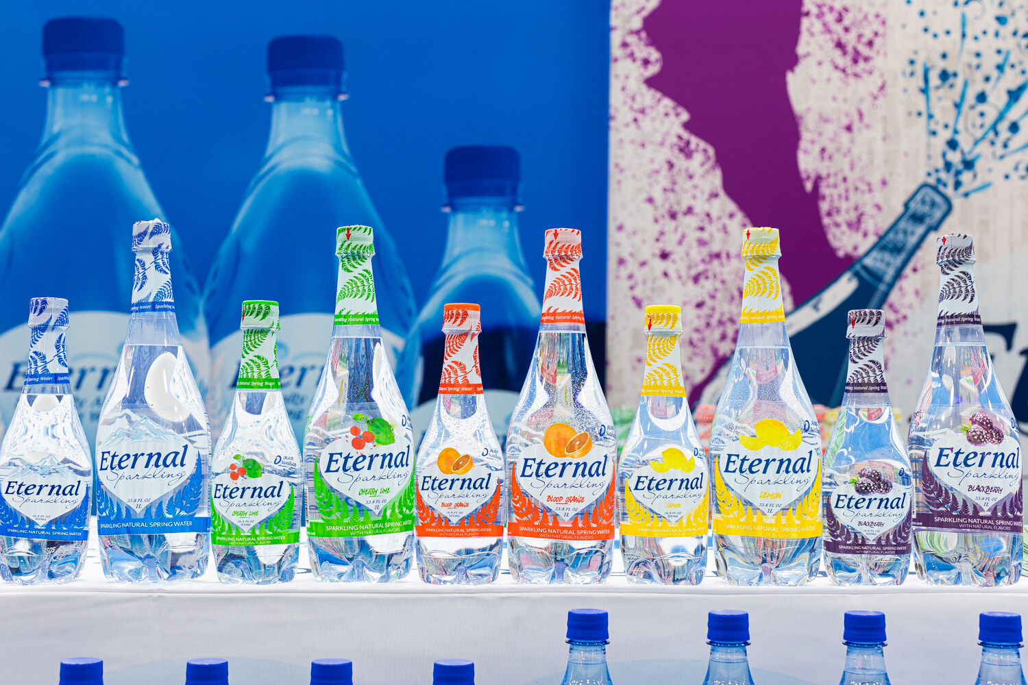 Bottles of Eternal sparkling water in blue, green, orange, yellow and purple labels on a backlit shelf.