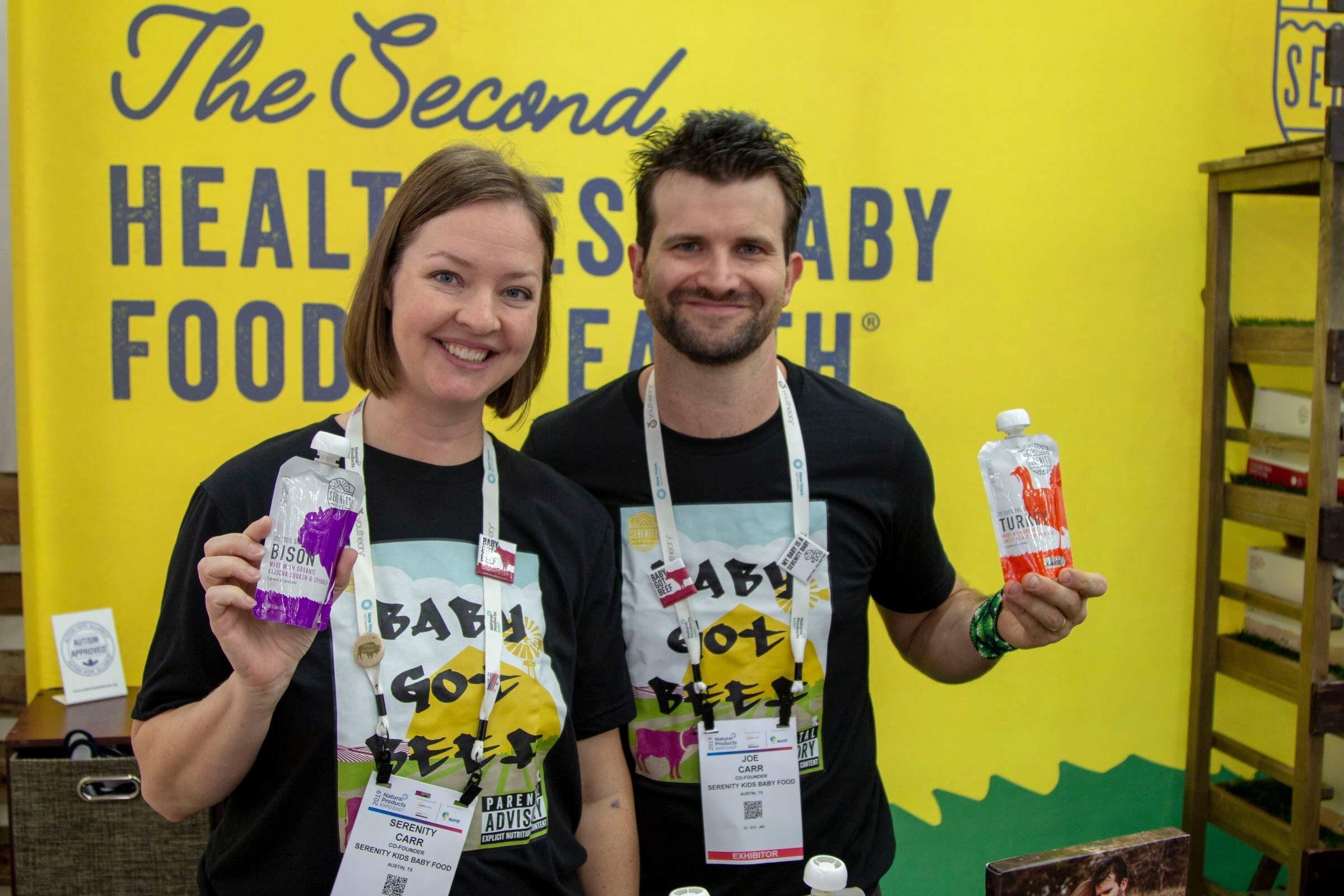 A woman and a man in black shirts and event badges holding small drink pouches in front of a yellow wall.