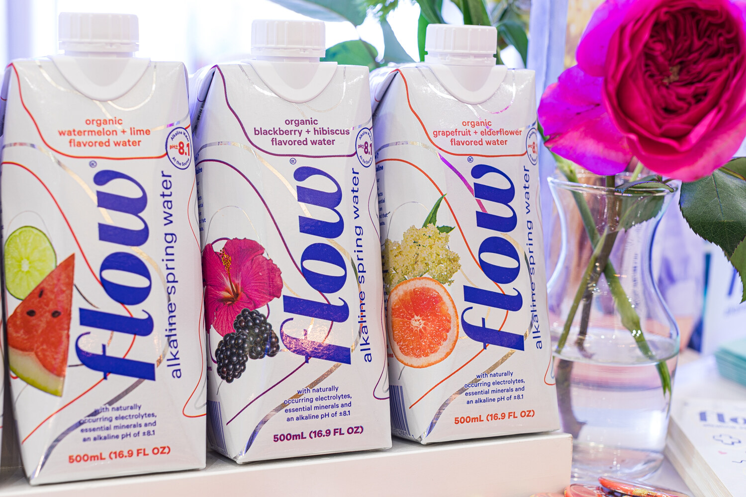 Flow alkaline spring water cartons in watermelon-lime, blackberry-hibiscus and grapefruit-elderflower, with a pink rose at right.