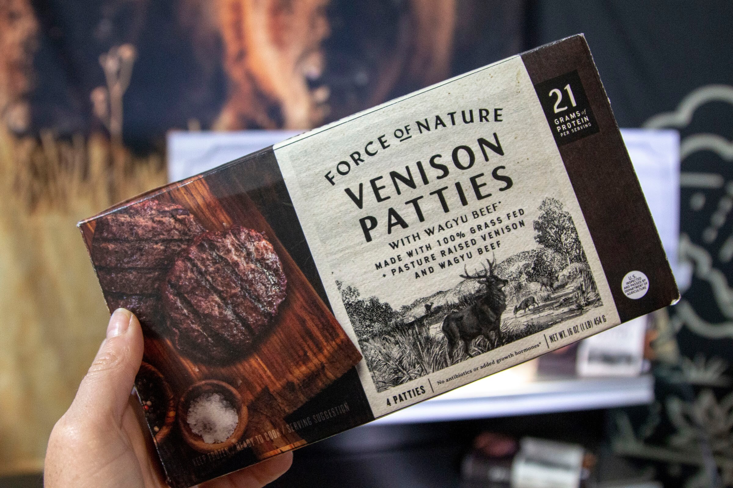 A hand holding a Force of Nature Venison Patties box in front of a dark booth backdrop.