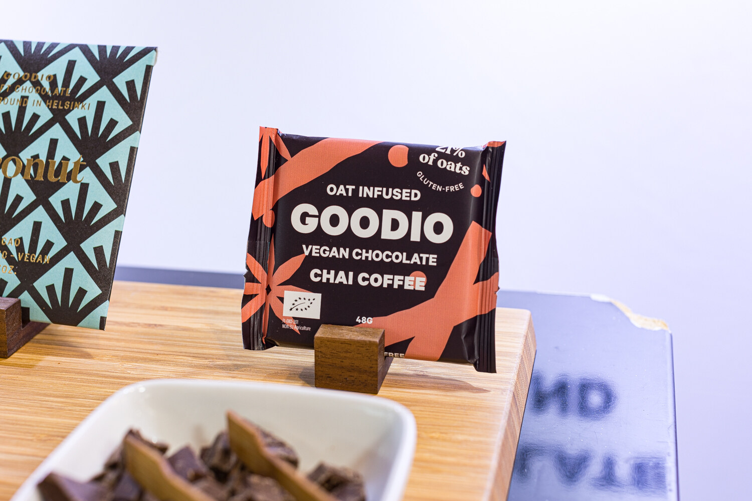 A Goodio vegan chocolate bar on a wooden stand with a bowl of chocolate pieces in the foreground.