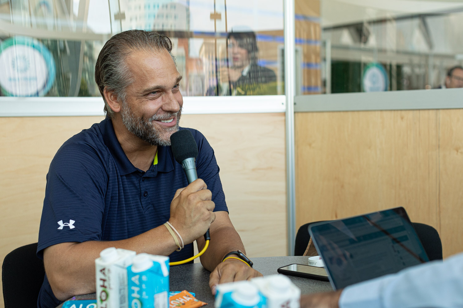A man in a navy polo holding a microphone at a table, smiling, with laptop and product cartons in front of him.