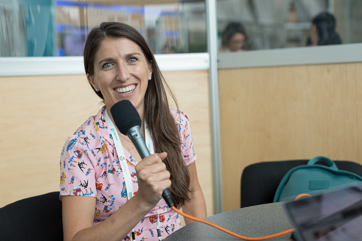 A woman in a pink floral dress holding a microphone, seated at a round table in a glass-walled booth.