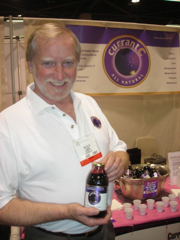 A smiling bearded man in a white polo holds a bottle of CurrantC juice in front of a purple CurrantC All Natural banner, with sample cups and bottles on a table behind him.