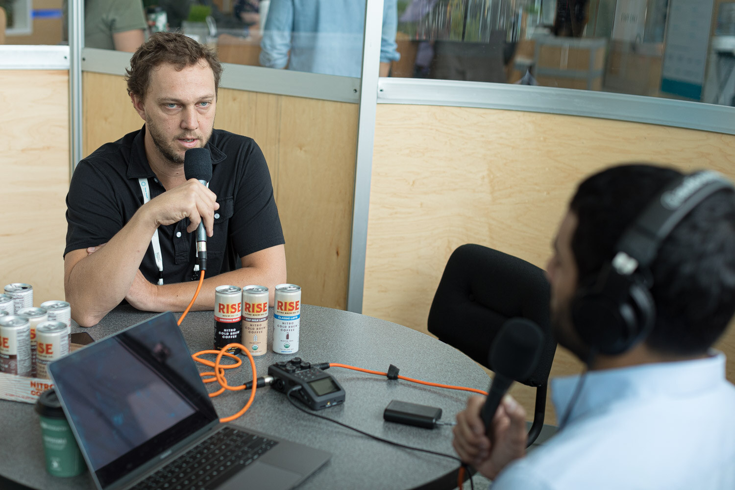 A man in a black shirt with a microphone at a round table across from a man wearing headphones.