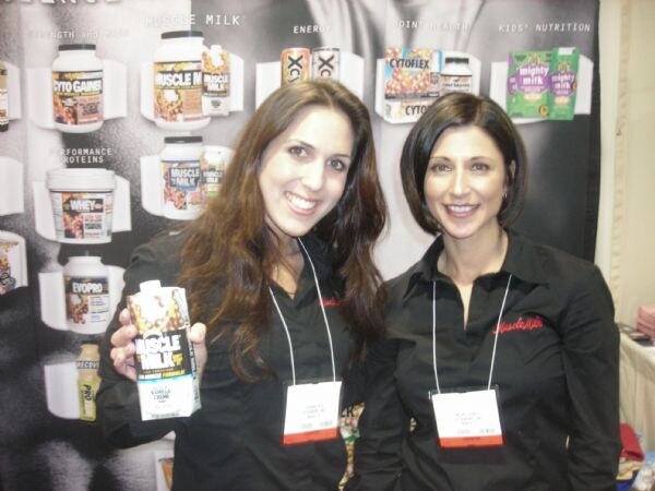 Two smiling women in black shirts stand before a display of Cytosport products including Muscle Milk, Cytogainer, Evopro, Cytoflex and Mighty Milk; one holds a Muscle Milk carton.