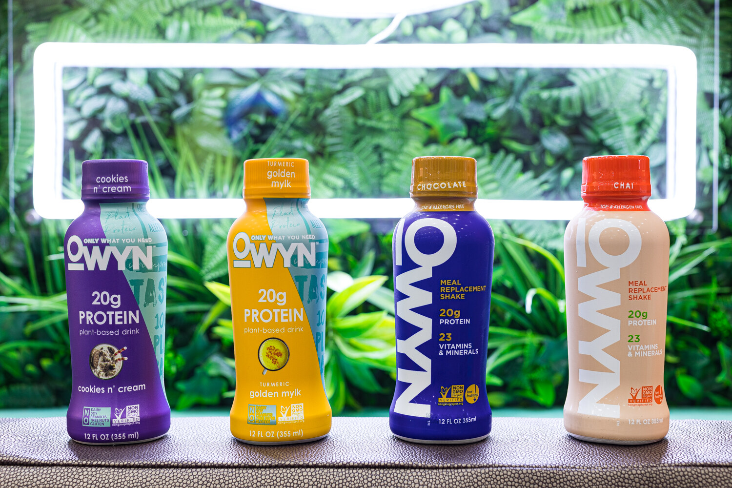 Bottles of OWYN protein drinks in cookies n' cream, golden milk, chocolate and chai flavors against a leafy backdrop.