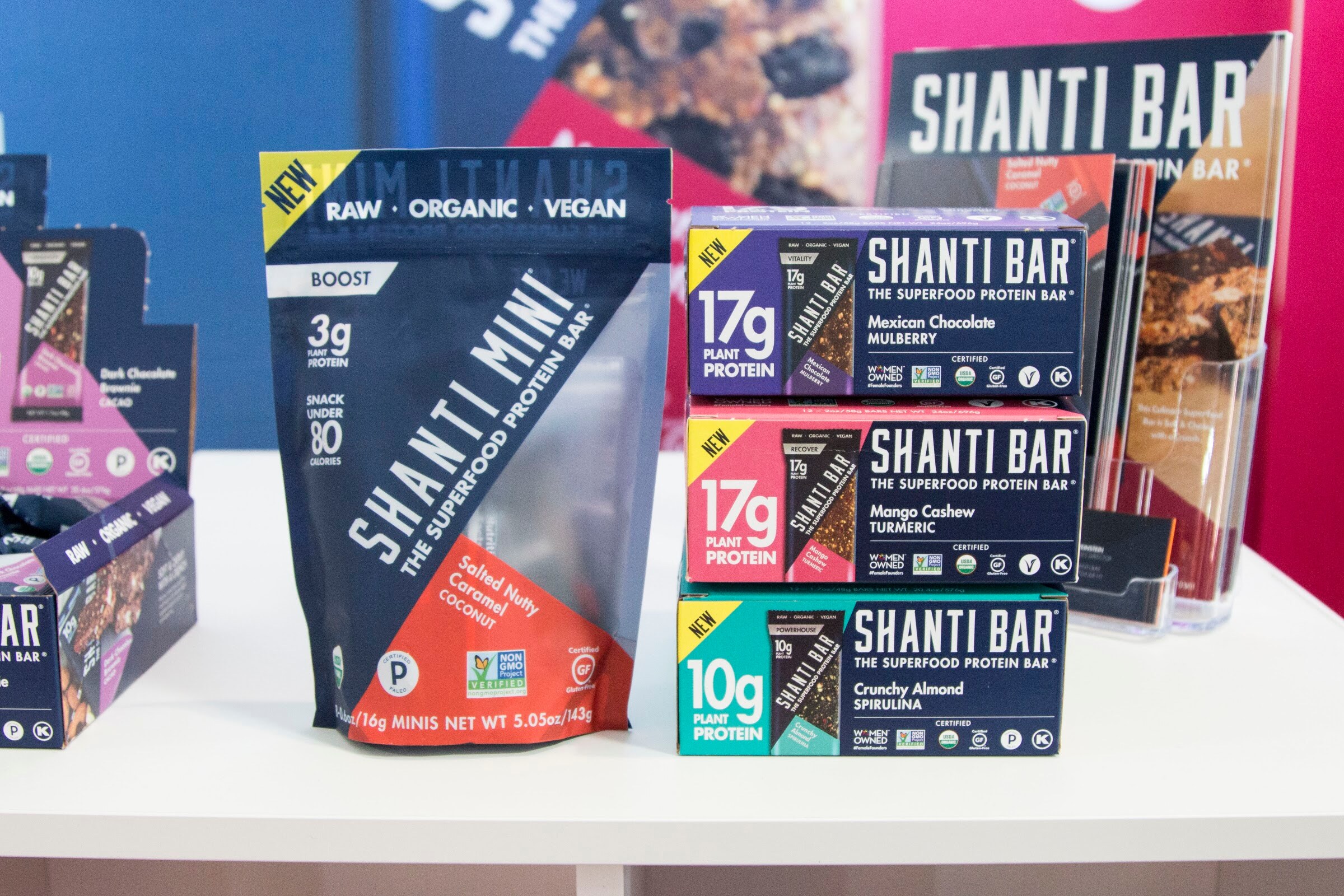 A Shanti Mini pouch and boxes of Shanti Bar protein bars on a white table.
