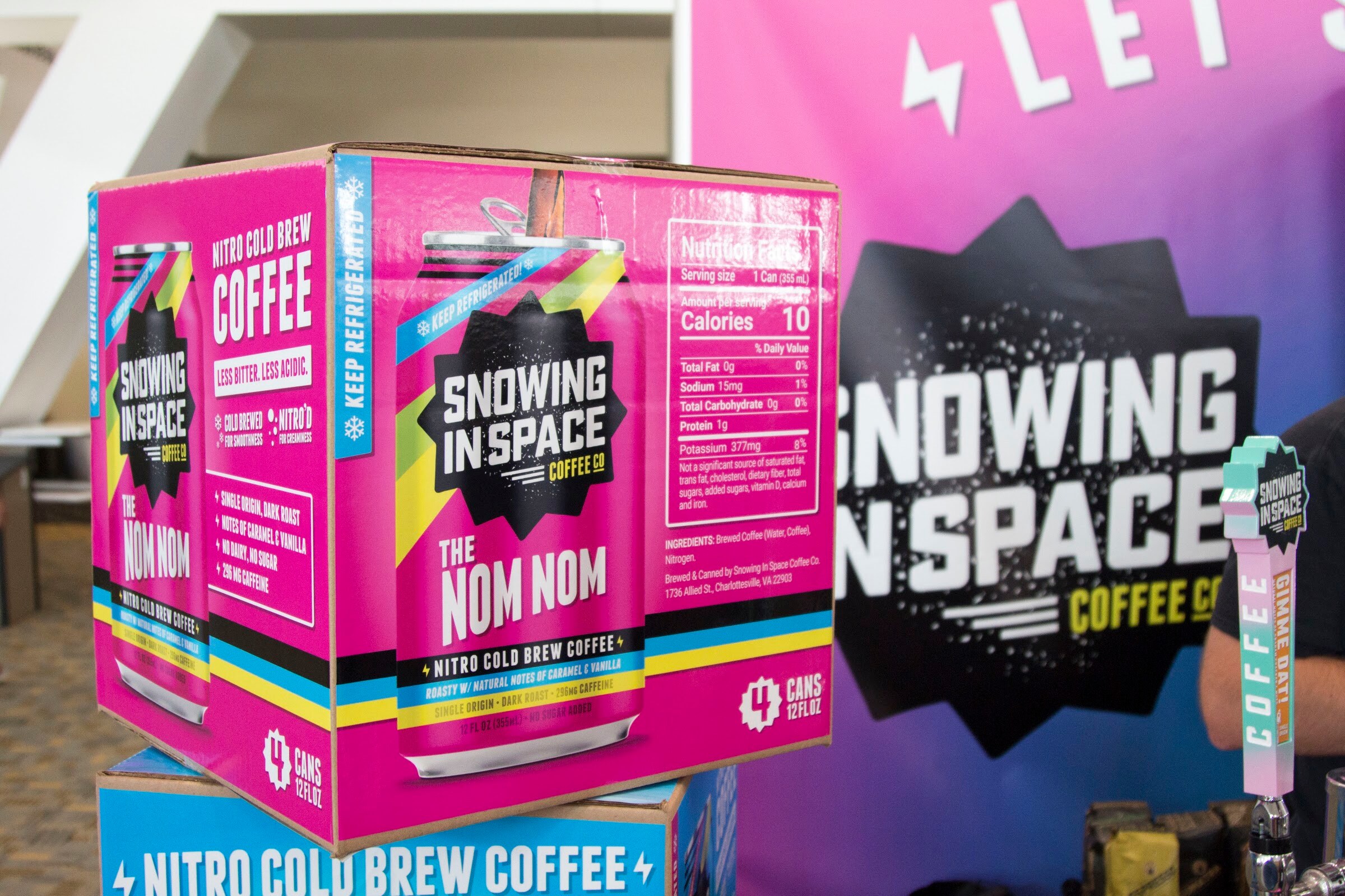 A pink and blue box of Snowing in Space The Nom Nom nitro cold brew coffee in front of a black Snowing in Space sign.