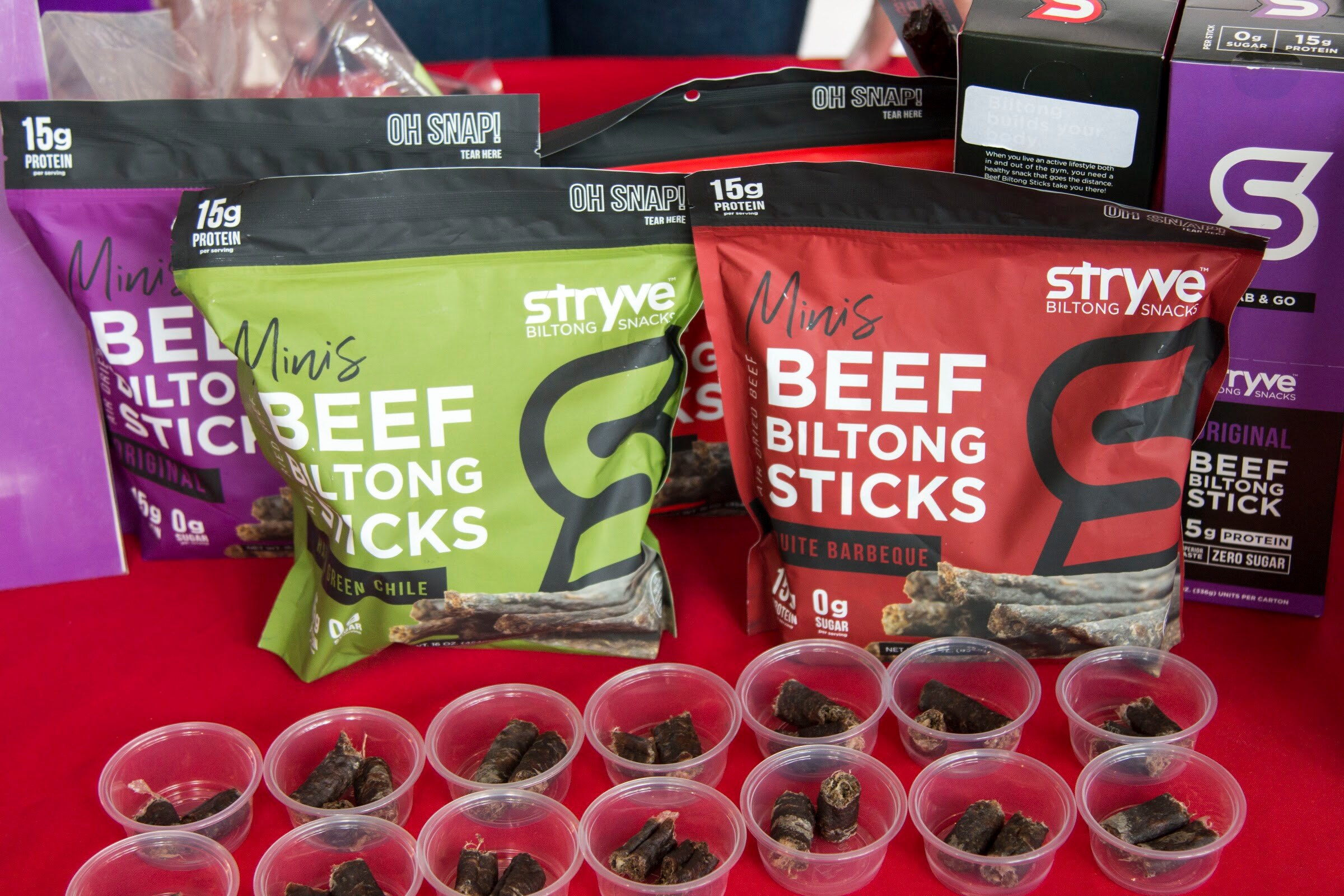 Bags of Stryve Mini's Beef Biltong Sticks with small tasting cups in front on a red table.