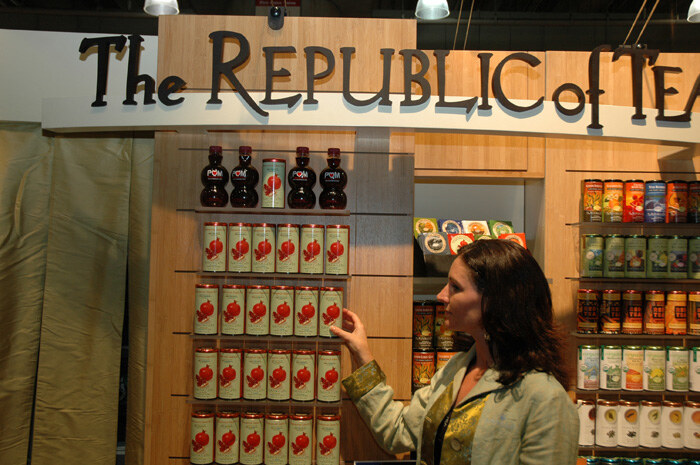 A woman in a green jacket reaches toward shelves of tea tins and POM bottles under a The Republic of Tea sign, with more tins on shelves at right.