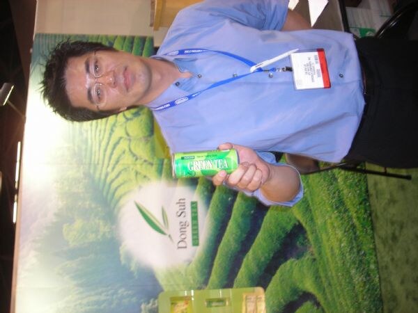 A man with glasses and a blue shirt holds a can of Green Tea in front of a Dong Suh banner showing green tea fields. The photo is rotated sideways.