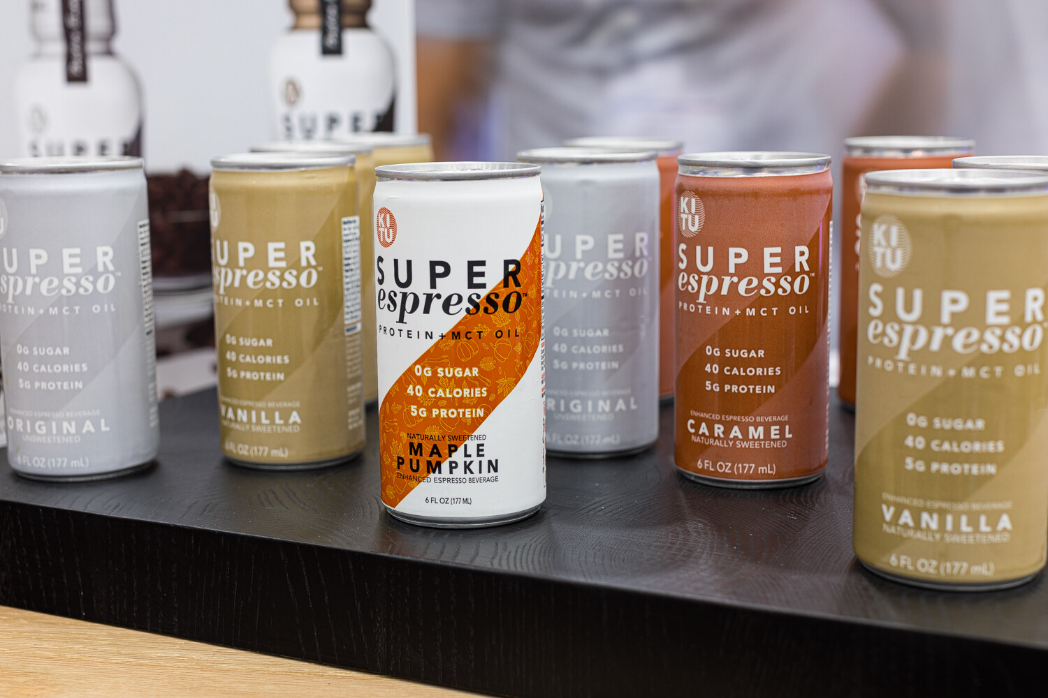 Small cans labeled Super Espresso in Original, Vanilla, Caramel and Maple Pumpkin on a black shelf.