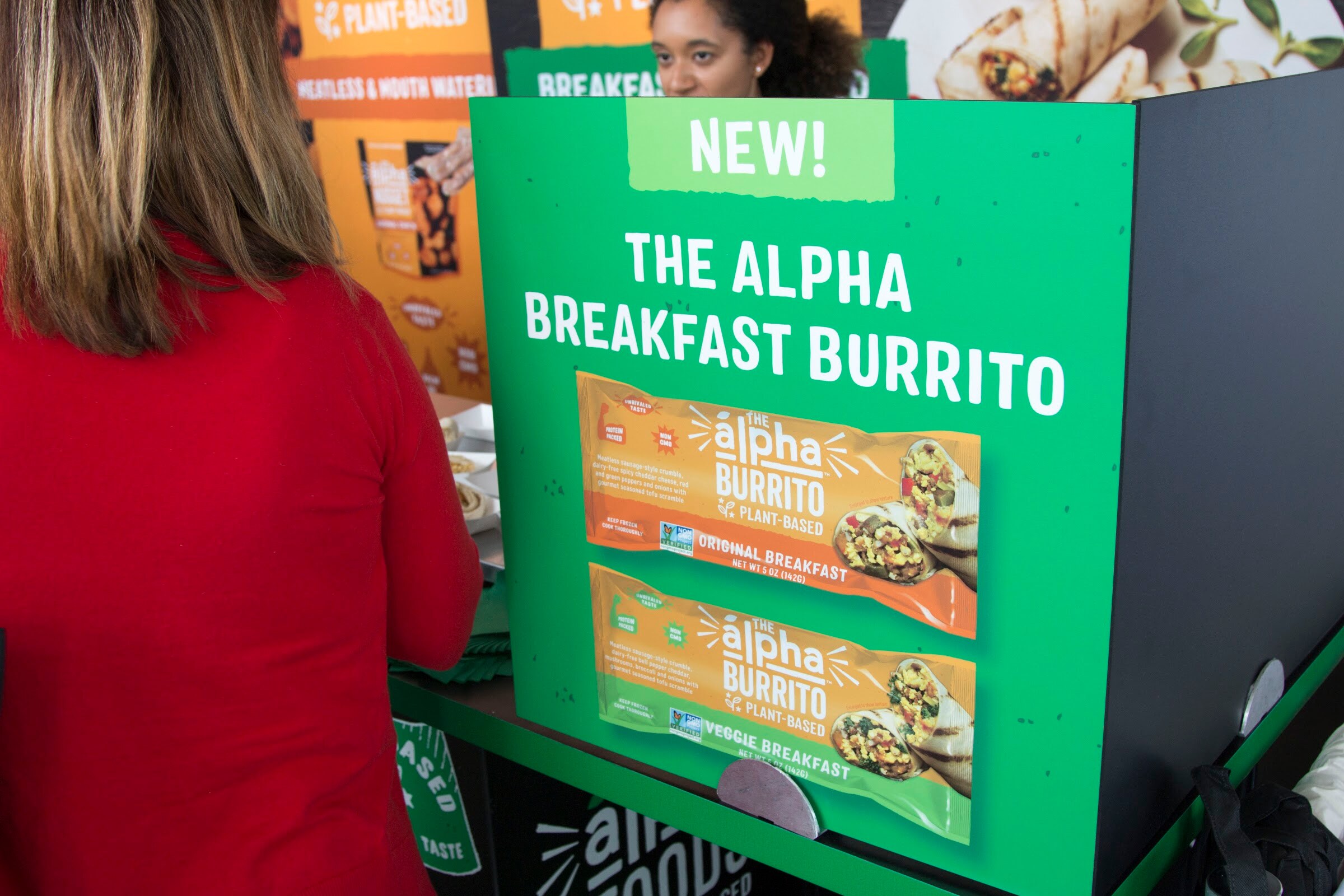 A green countertop sign reading NEW! The Alpha Breakfast Burrito, with burritos displayed below and a visitor in red at left.