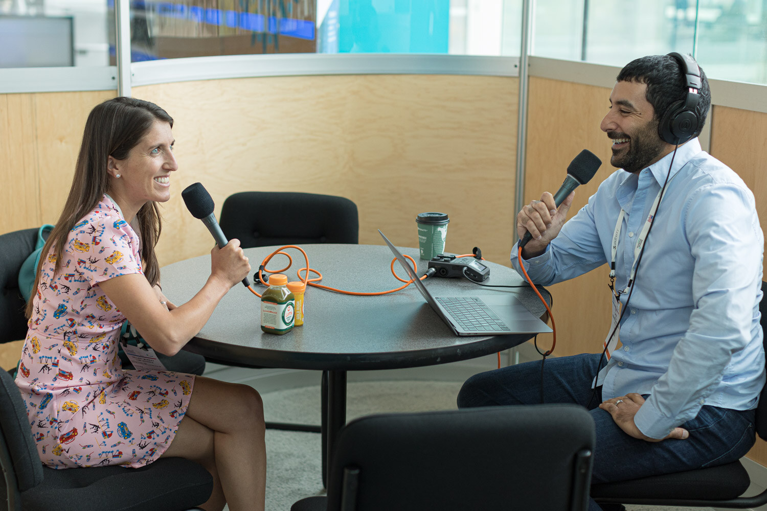 A woman in a floral dress and a man in a blue shirt wearing headphones, each holding a microphone, laughing at a table.