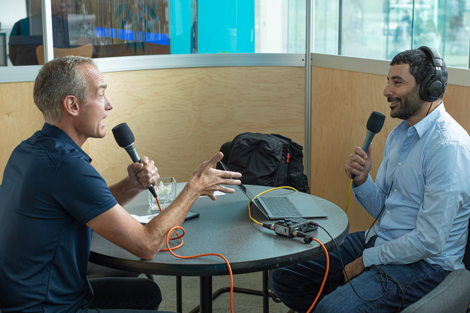 A man in a navy shirt gestures while speaking into a microphone across from a man in a blue shirt wearing headphones.
