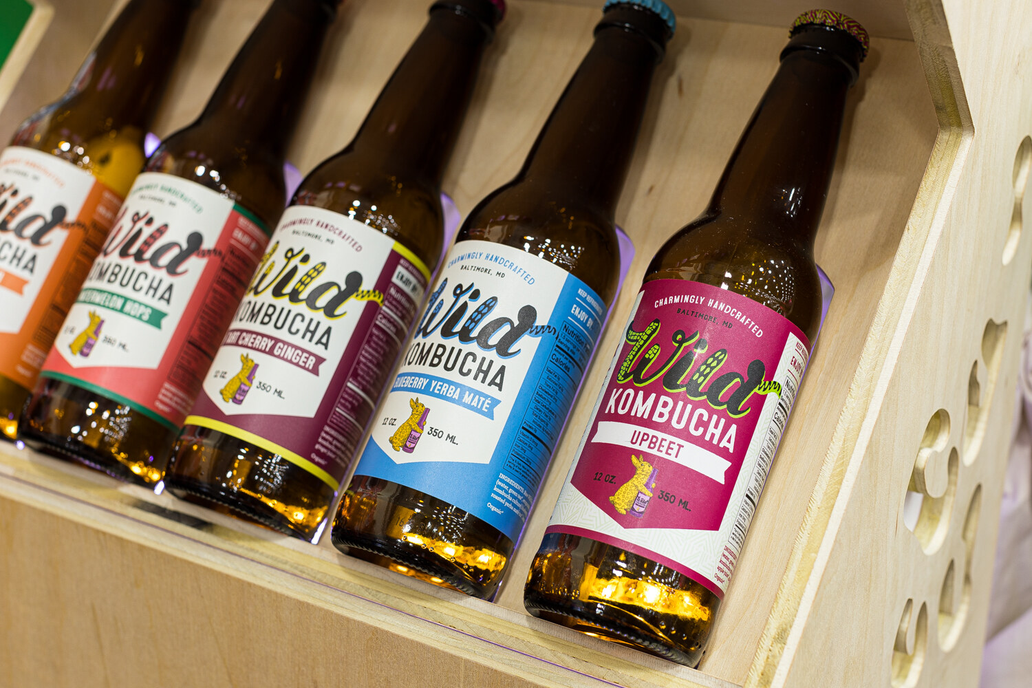 Five bottles of Wild Kombucha with colorful labels resting in an angled wooden rack.