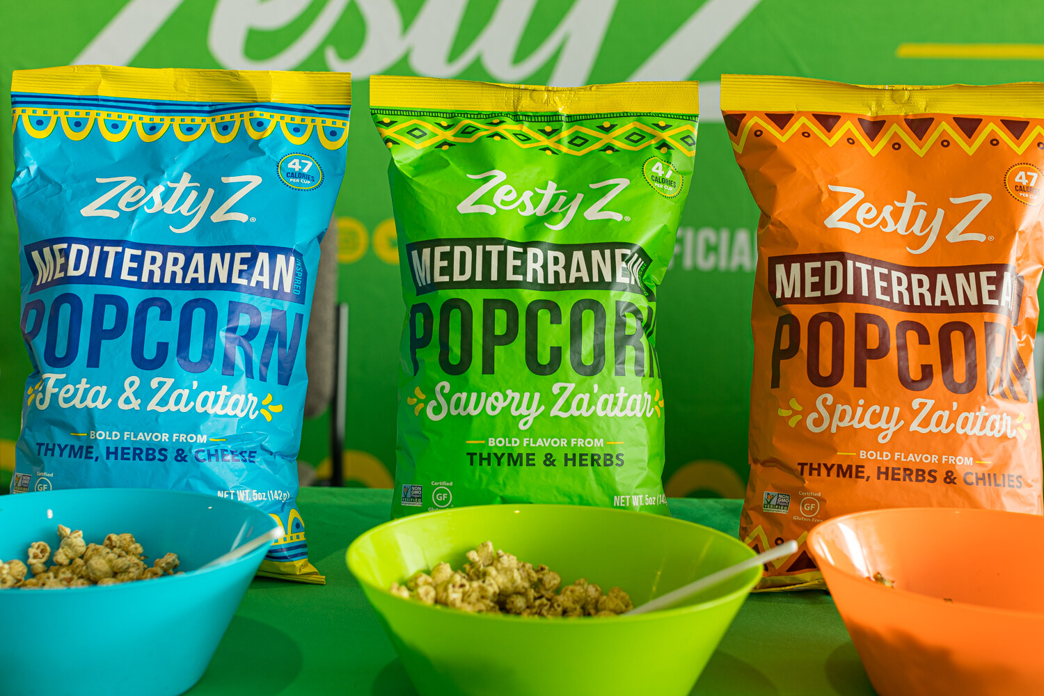 Three Zesty Z popcorn bags in feta and za'atar, savory za'atar and spicy za'atar flavors, with green and orange sample bowls.
