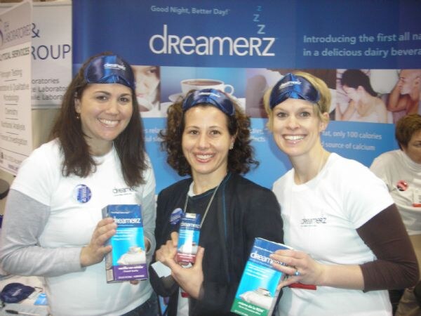 Three smiling women in white shirts and blue sleep masks on their foreheads hold Dreamerz cartons in front of a blue Dreamerz banner.