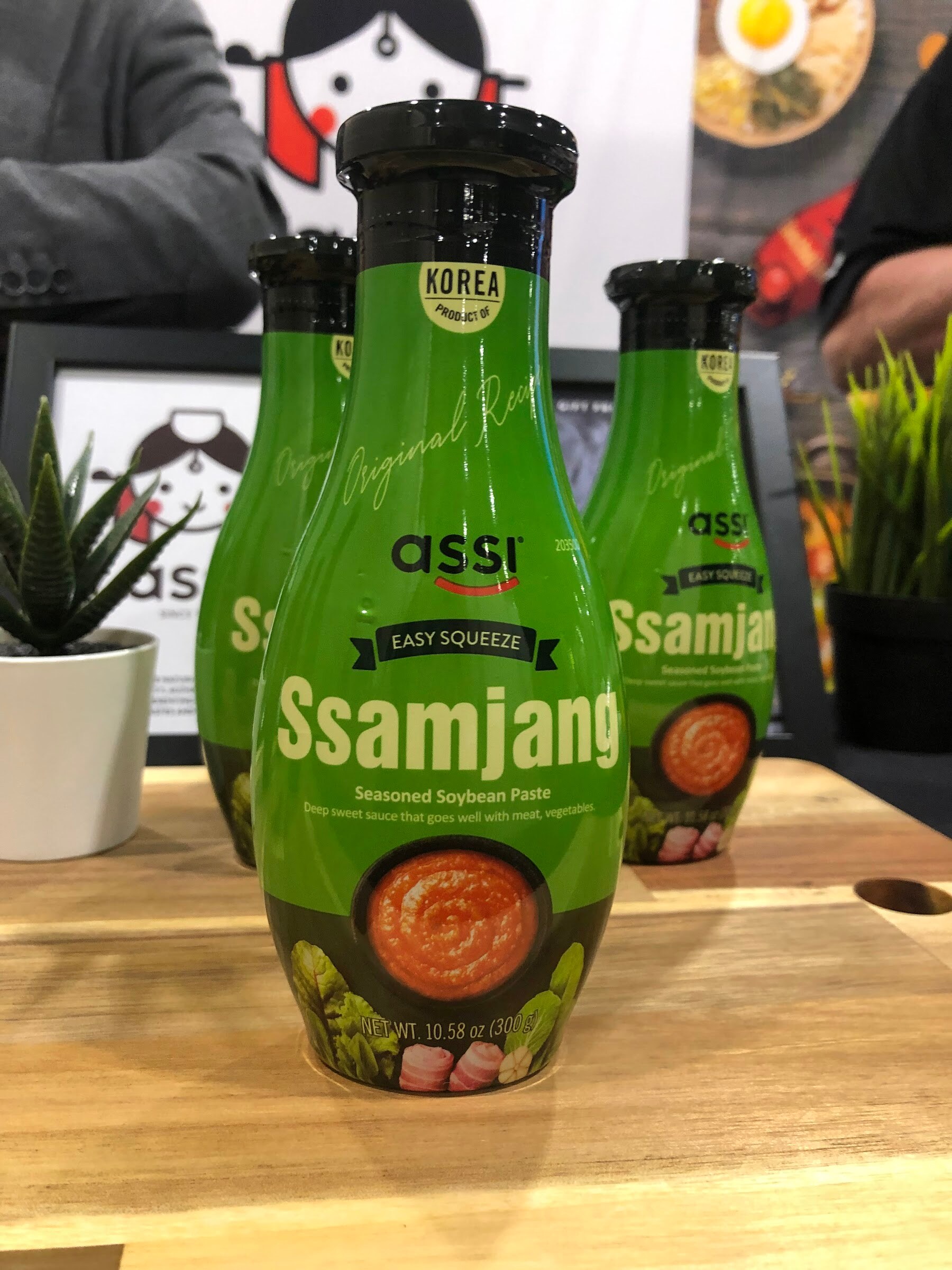 Green assi Easy Squeeze Ssamjang seasoned soybean paste bottles on a wooden board with a small plant behind