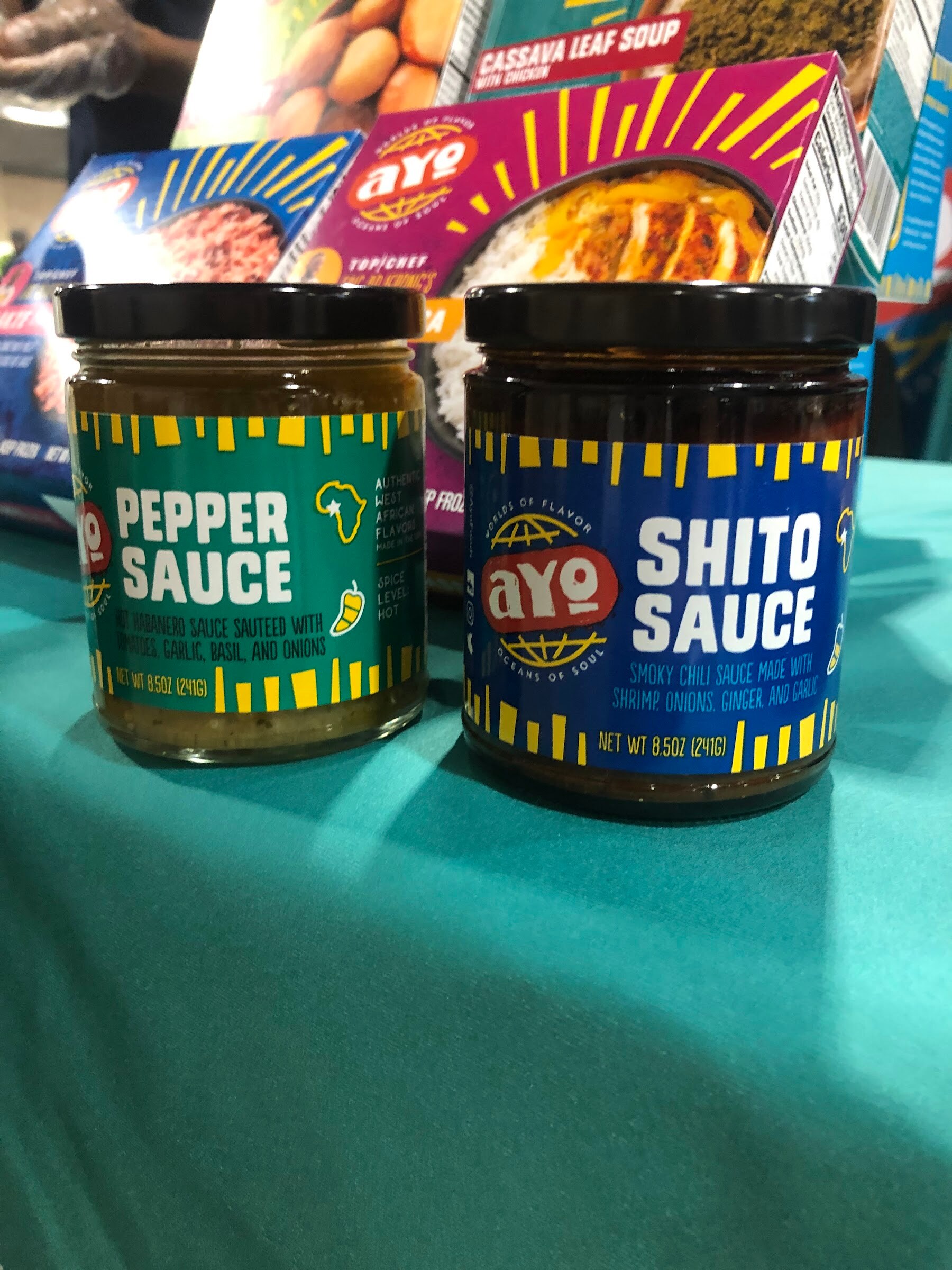 Jars of AYO Pepper Sauce and Shito Sauce on a teal tablecloth with AYO boxed meals behind them