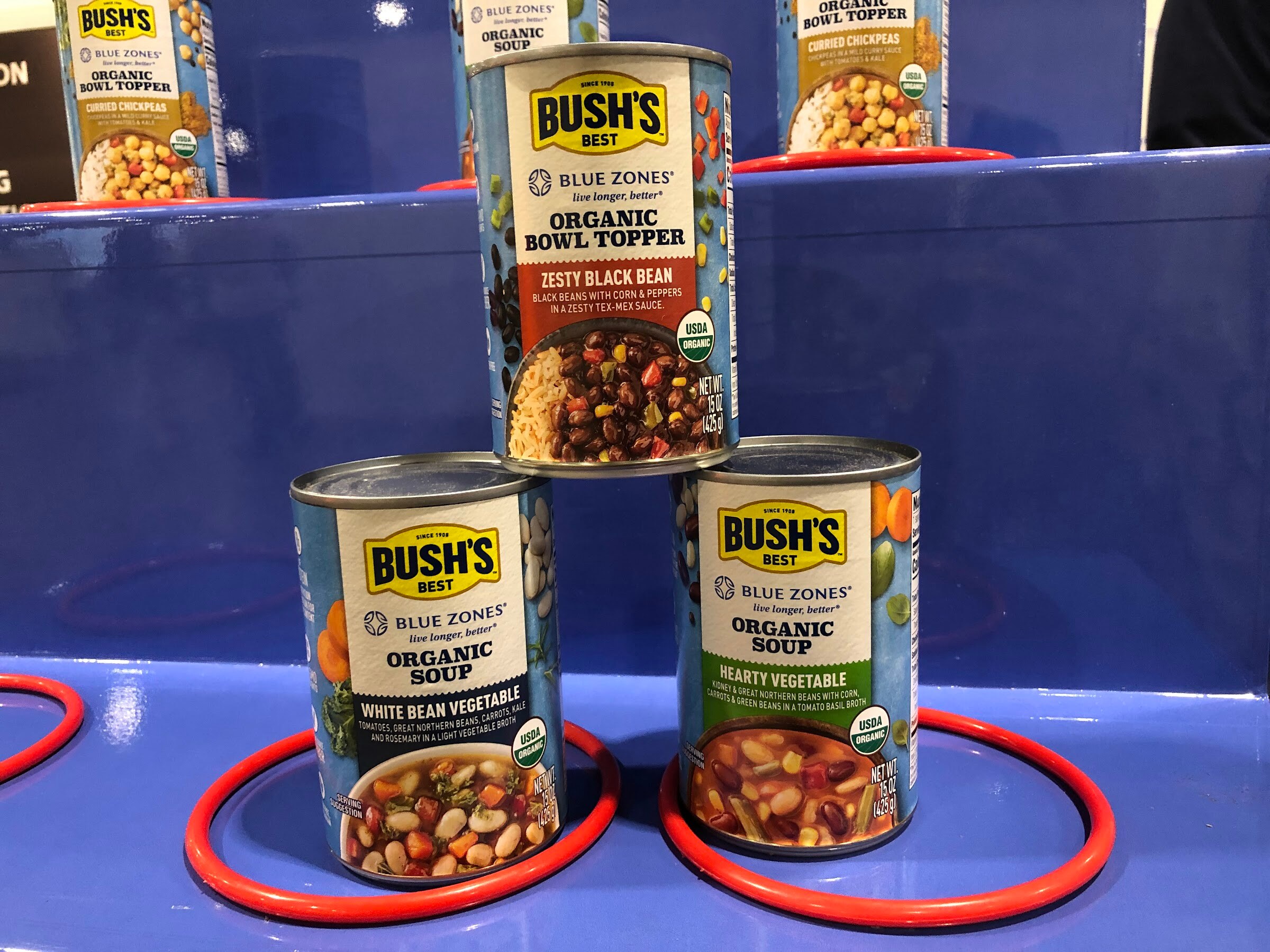 A pyramid of Bush's Best cans with Blue Zones organic bowl topper and soups on a blue stand with red rings