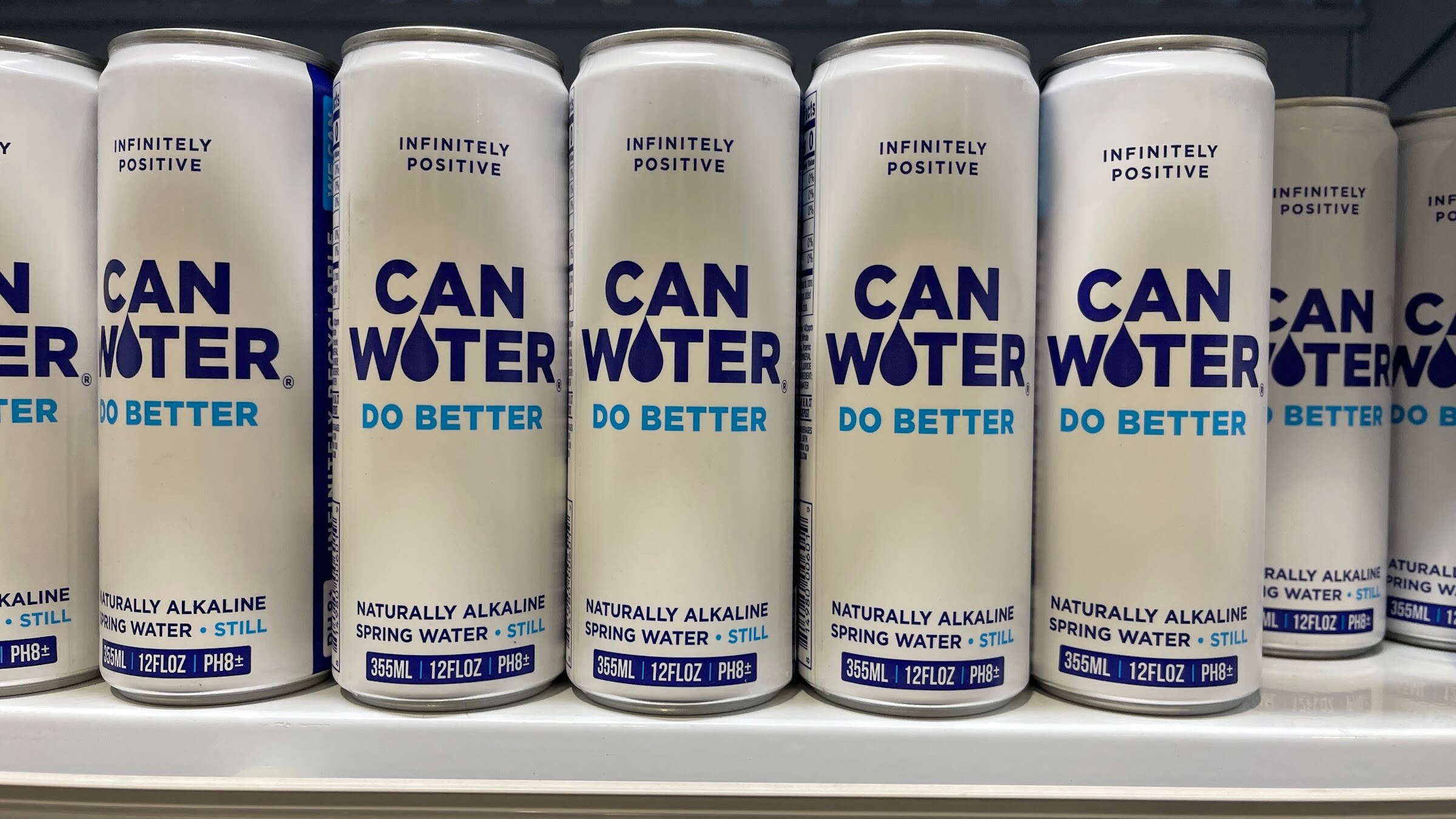 Six CAN WATER alkaline spring water cans with Do Better printed on them stand in a row on a white shelf
