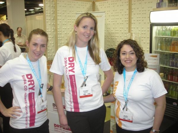 Three smiling women in white DRY Soda Co. T-shirts and blue lanyards stand in a booth, with a refrigerated case of bottles on the right.