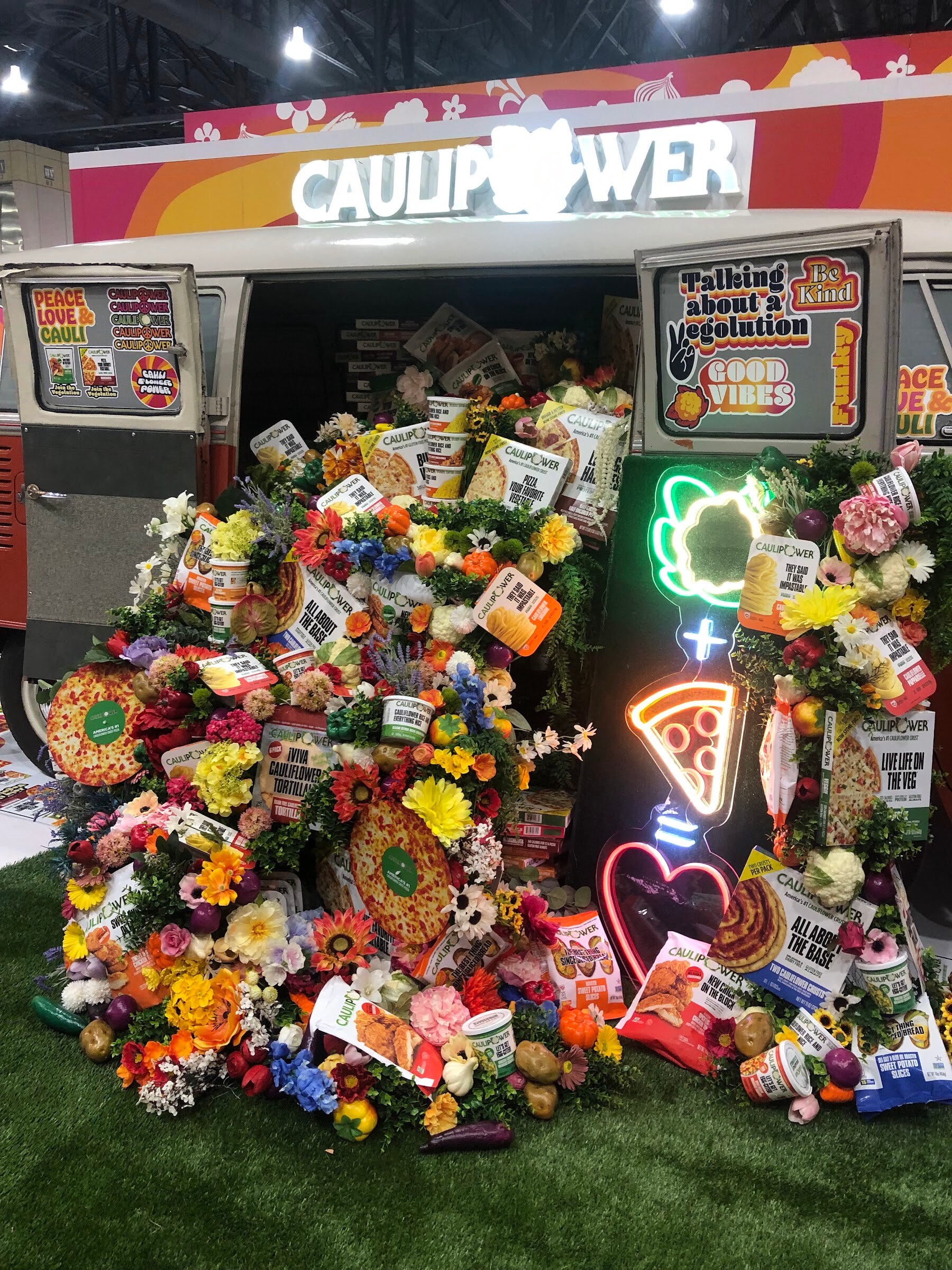A van-shaped Caulipower booth covered in flowers and product packages, with neon vegetable, pizza and heart signs
