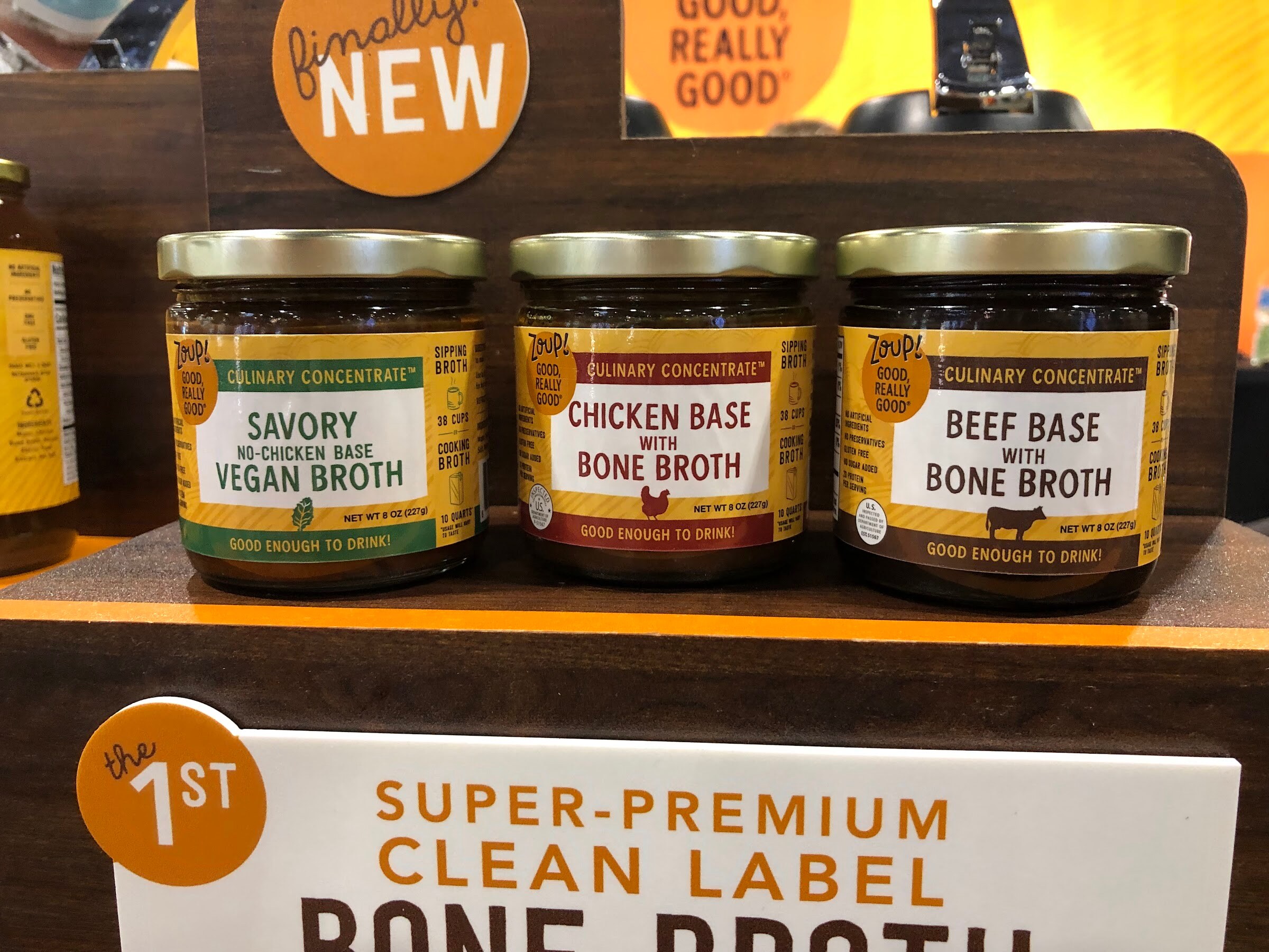 Three jars of culinary concentrate, vegan broth, chicken bone broth and beef bone broth, on a wooden stand with a 1st clean label bone broth sign