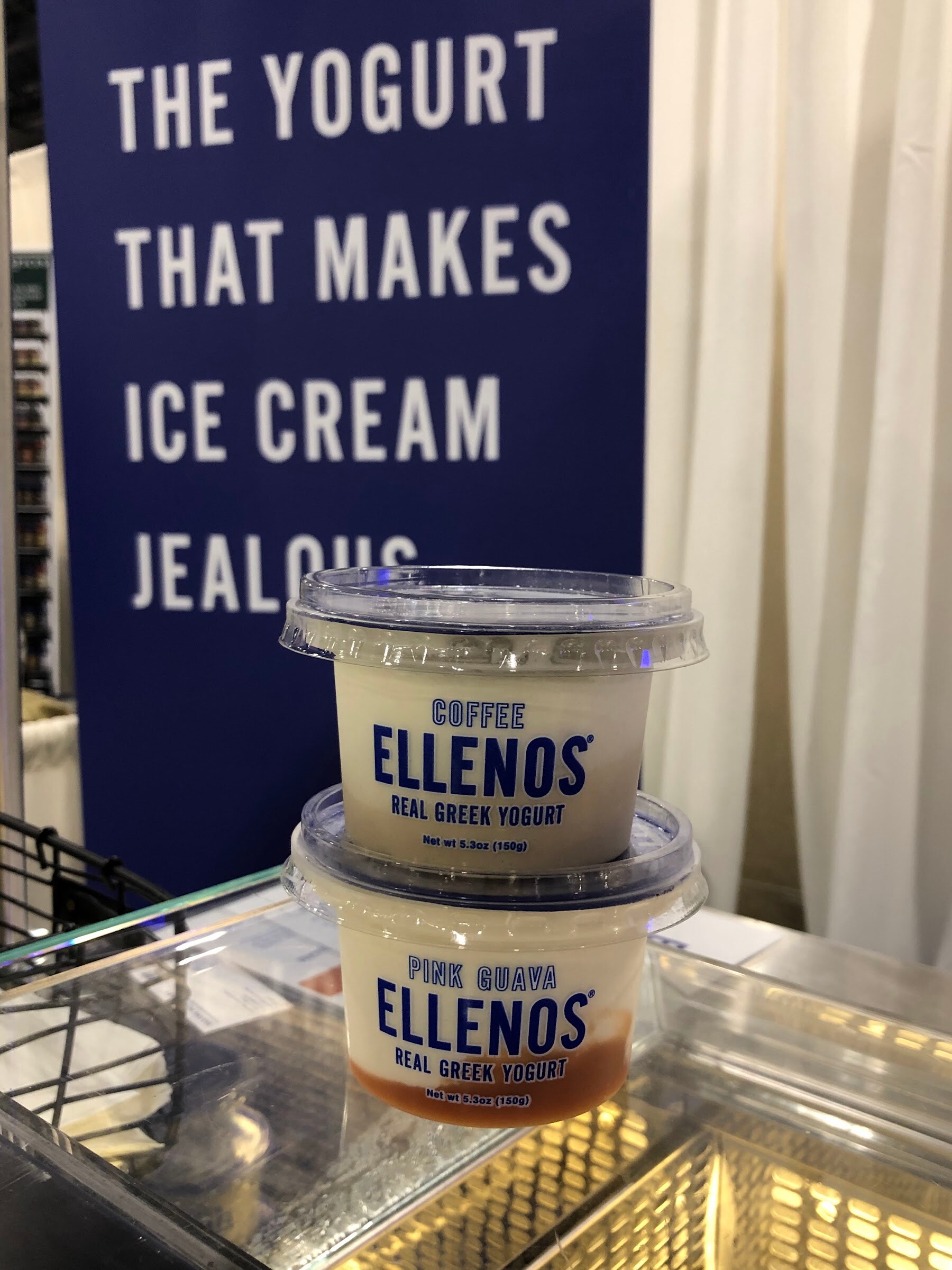 Two stacked Ellenos real Greek yogurt cups, coffee and pink guava, in front of a blue sign about ice cream jealousy