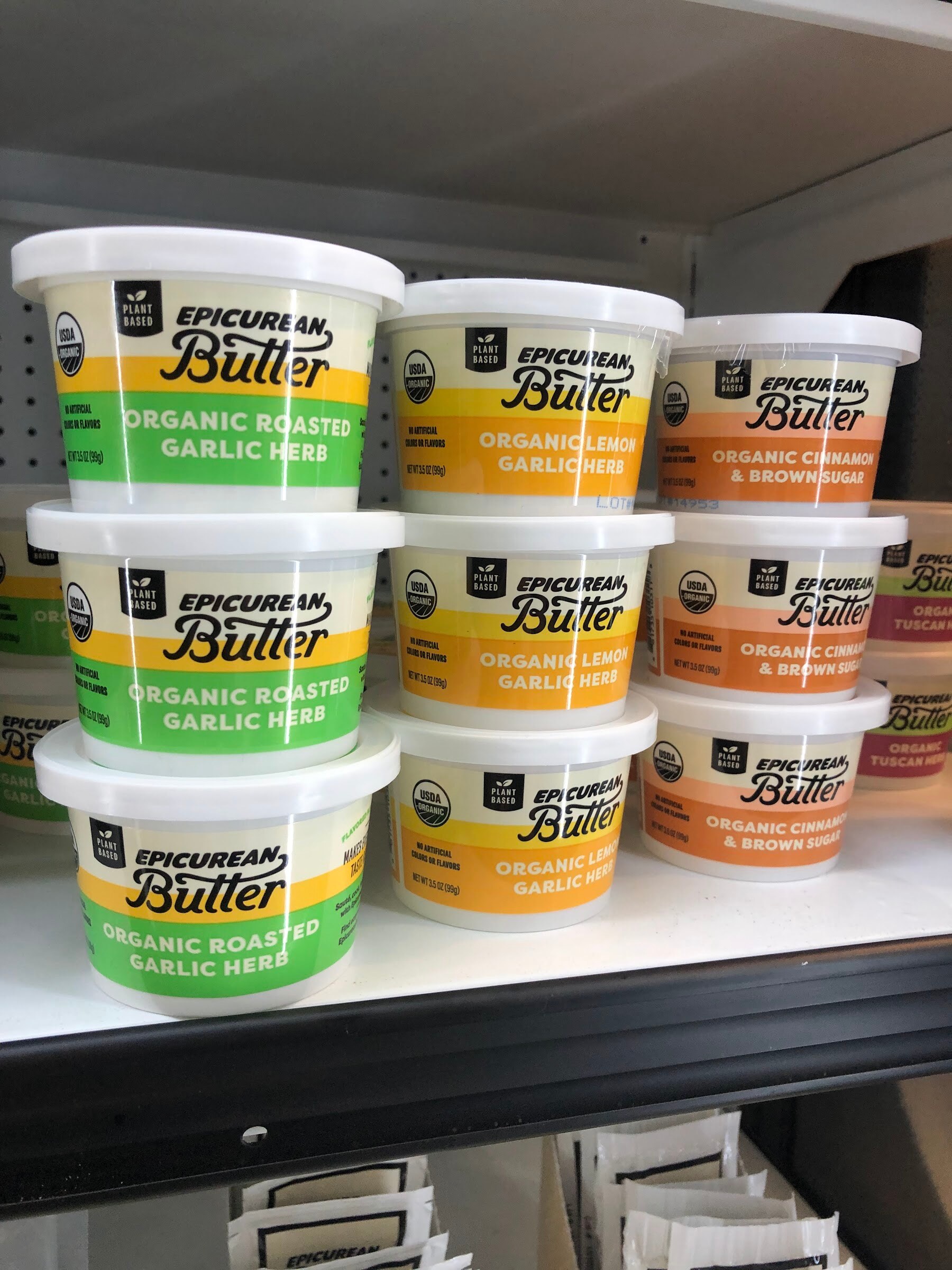 Stacked tubs of Epicurean Butter in organic roasted garlic herb and cinnamon brown sugar flavors on a refrigerated shelf