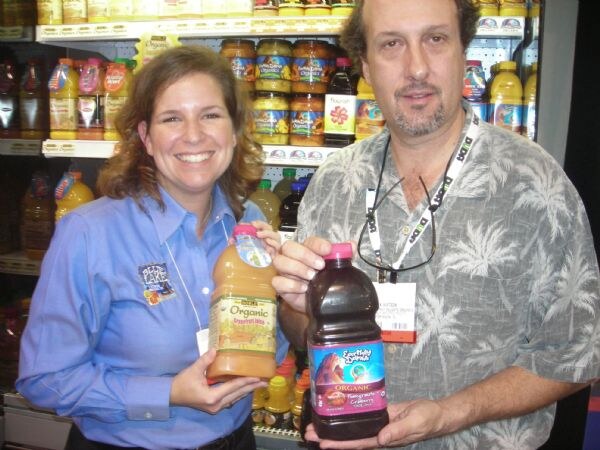 A smiling woman in a blue shirt and a man in a palm-print shirt hold bottles of Earthly Delights organic juice in front of shelves of bottled juices and jarred goods.