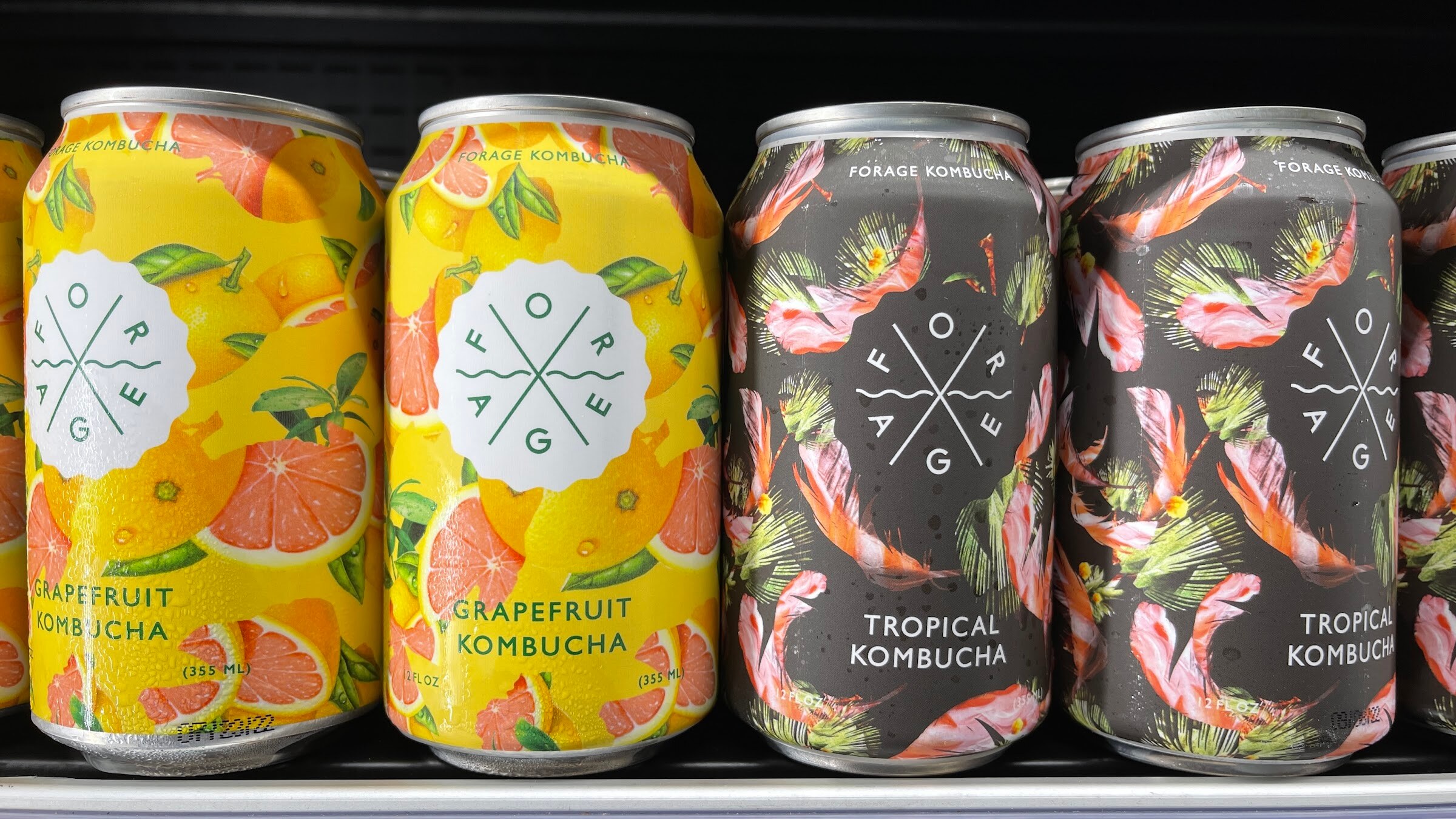 Yellow Forage grapefruit kombucha cans and dark floral tropical kombucha cans in a row on a shelf