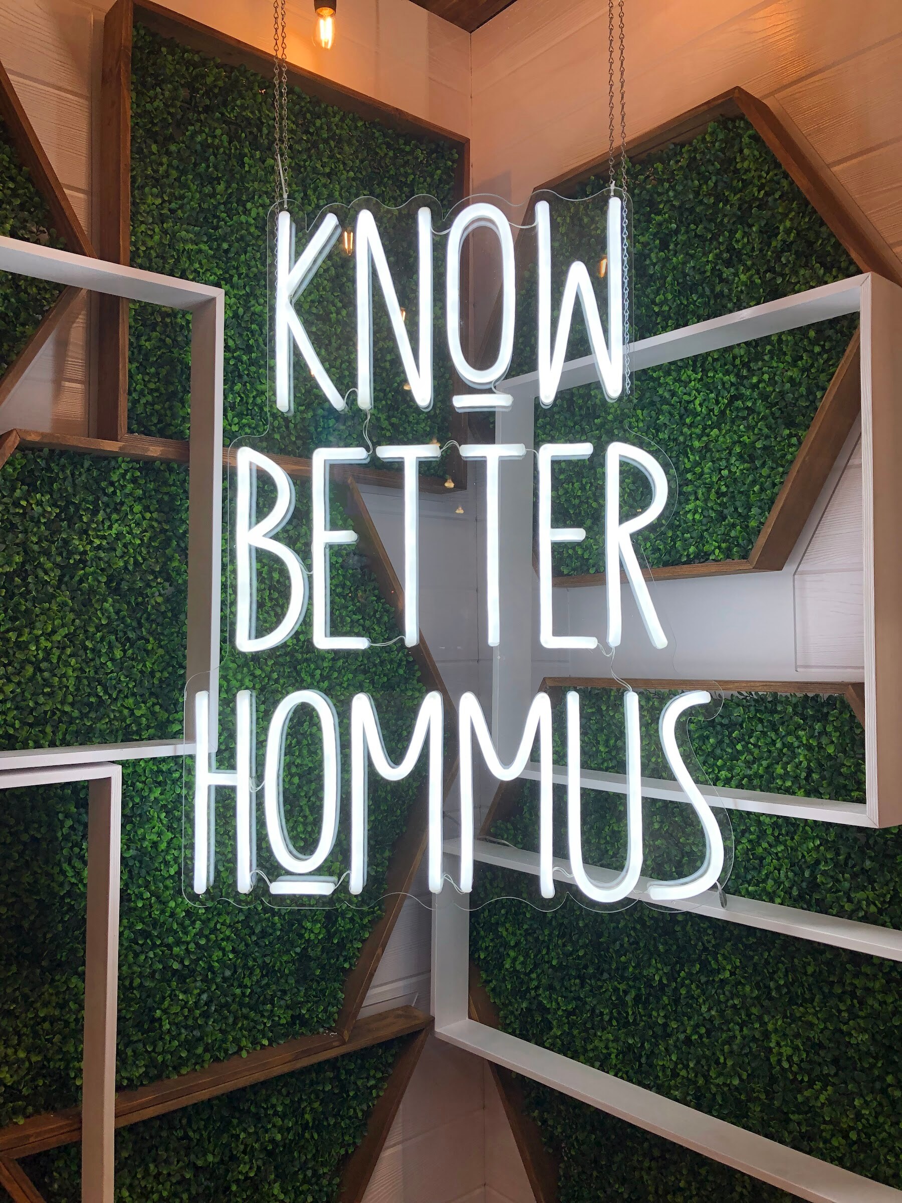 A white neon sign reading Know Better Hummus on a hedge-style wall