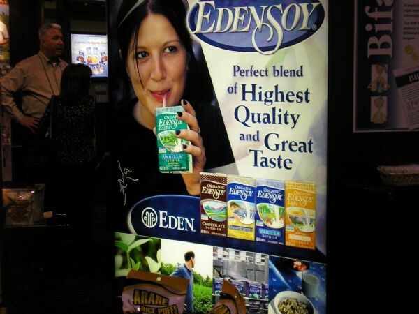 A lit EdenSoy display shows a woman drinking from a carton, above cartons of chocolate, vanilla and other flavors, with an Eden logo. A man stands in the background.
