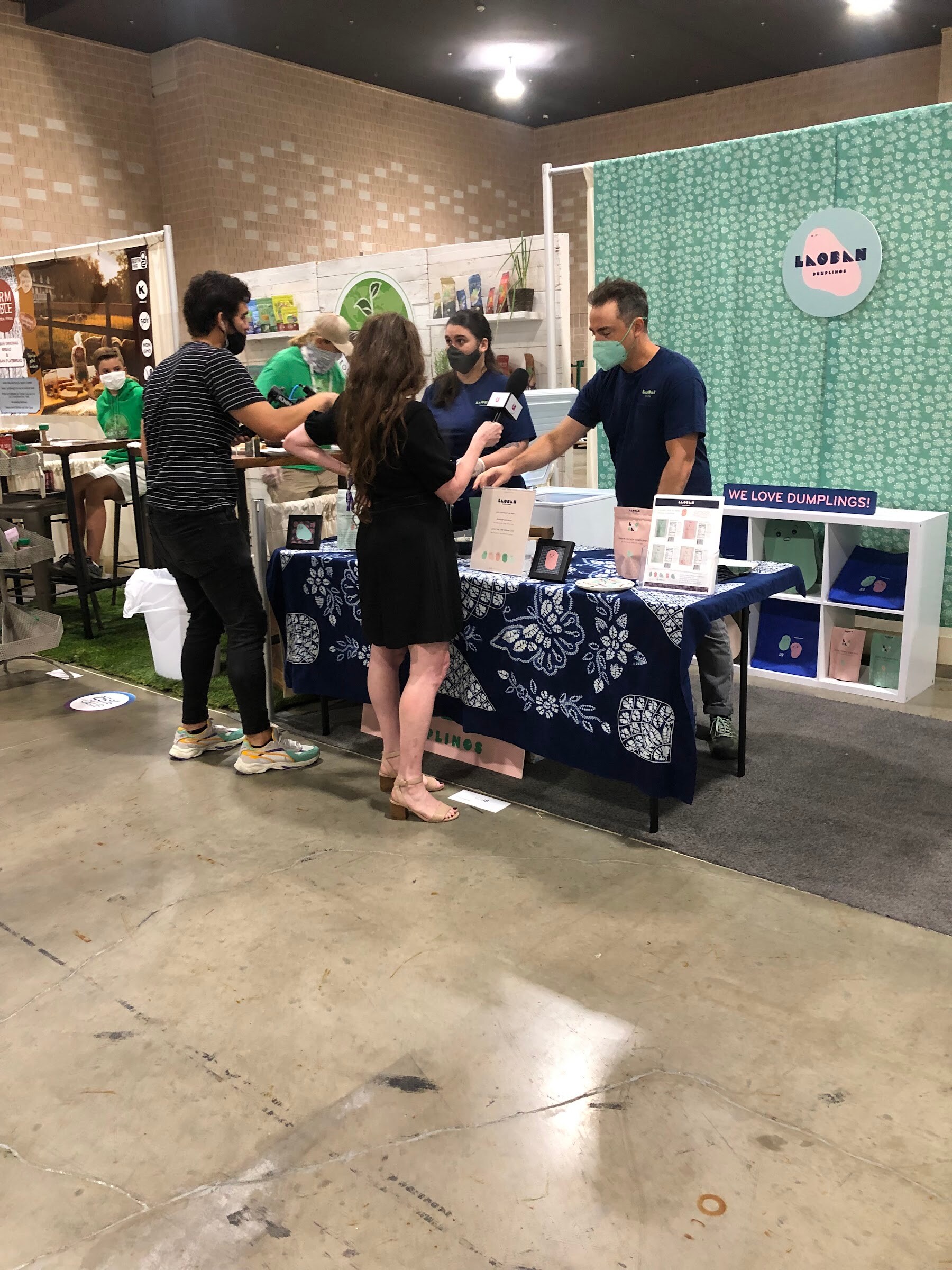 Exhibitors and visitors talk at a Laoban Dumplings booth with a blue patterned table and teal backdrop on the show floor
