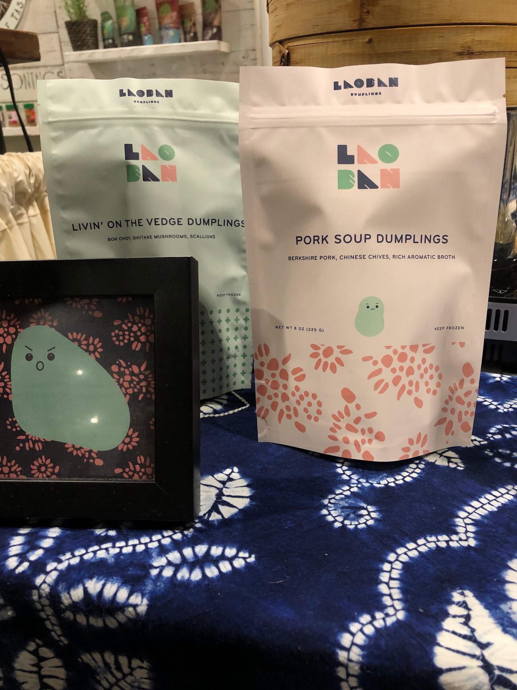 Laoban Dumplings pork soup and vegetable dumpling pouches beside a framed illustration on a blue patterned cloth
