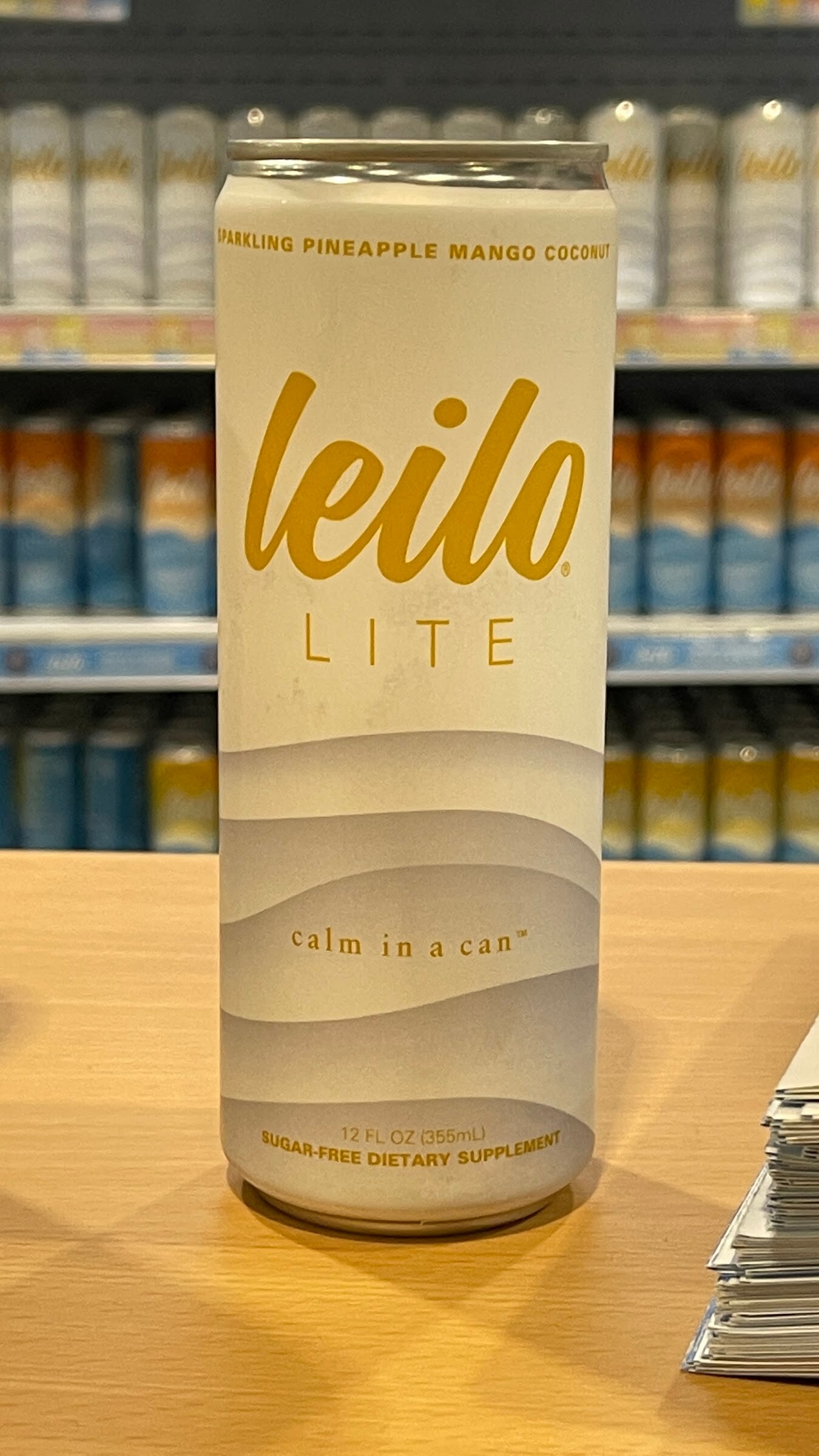 A white leilo Lite sparkling pineapple mango coconut can on a wooden table with shelves of cans behind it